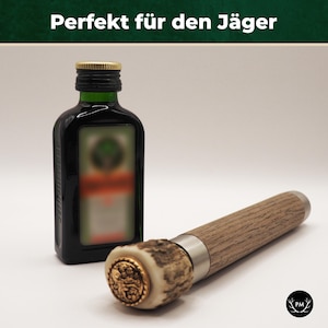 Könnte beinhalten: Ein Flaschenverschluss aus Holz mit einem Metallband und einer goldfarbenen Verzierung. Der Verschluss liegt neben einer kleinen Flasche mit dunkler Flüssigkeit und einem grünen Etikett. Das Etikett trägt den Text "Jägermeister".