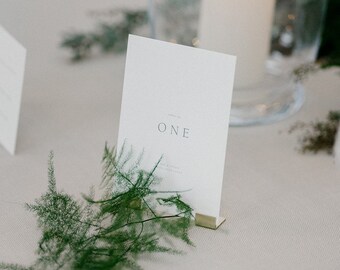 May include: A white card with the text "Table No. ONE" on it, sitting on a table with a green plant and a candle in a glass holder.