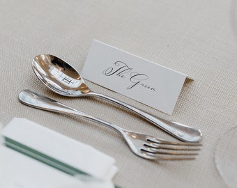 May include: A white place card with the text "The Groom" in black calligraphy, placed on a beige tablecloth with a silver spoon and fork.