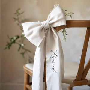 Op de afbeelding: Een witte stoffen strik met rafelige randen, gebonden om de rugleuning van een houten stoel. De stof heeft het woord "Gereserveerd" in zwarte kalligrafie.