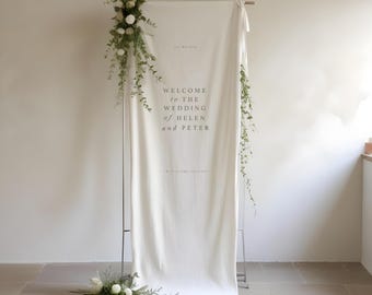 Könnte beinhalten: Weißer Hochzeitshintergrund mit Grünpflanzen und Blumen. Der Hintergrund hat den Text "Willkommen zur Hochzeit von Helen und Peter".