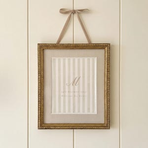 May include: A rectangular gold frame with a beaded edge containing a vertical striped fabric insert. The centre features the letter M, the date 24th December 2023, and the name Millicent Joy Caven. A beige ribbon is tied in a bow at the top.