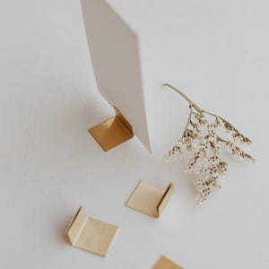 May include: Three small, L-shaped brushed brass metal stands for displaying cards, with one stand holding a blank white rectangular card. A small sprig of dried white flowers lies next to the metal stands on a plain white surface.