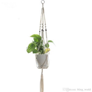 Puede incluir: Un macetero de macramé blanco con una maceta blanca y una planta verde. El macetero tiene un acento de cuentas de madera y una borla larga.