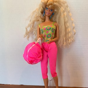 May include: A blonde Barbie doll wearing a green and floral print tank top, pink pants, and blue earrings. She is holding a pink bag and a pink hairbrush is in the foreground.