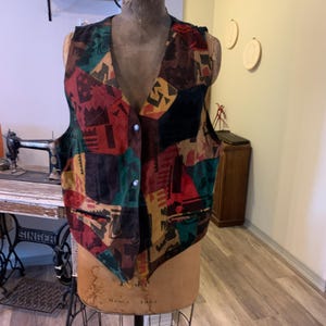 Könnte beinhalten: Eine Vintage-Weste mit Patchwork-Design in Rot-, Grün-, Braun- und Schwarztönen. Die Weste hat einen V-Ausschnitt und ist mit Knöpfen versehen. Die Weste wird auf einer Schaufensterpuppe präsentiert.