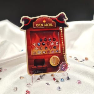 May include: A novelty acrylic charm featuring an "OVEN GACHA" design. The charm is shaped like a red oven with a witch hat on top, a gold coin, and a chain. The charm has a clear stand and is decorated with small, colorful gems.