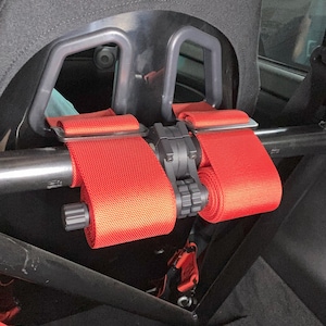 May include: Red racing harnesses are rolled up and secured to a black metal bar in the back of a car. The harnesses are attached to a black Sparco racing seat.