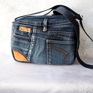 Könnte beinhalten: Eine Umhängetasche aus dunkelblauem Jeansstoff mit mehreren Taschen, einem beigefarbenen Ledereckaufnäher, einem braunen Lederetikett mit der Aufschrift "LS.D", das mit Metallnieten befestigt ist, und einem verstellbaren schwarzen Schultergurt.