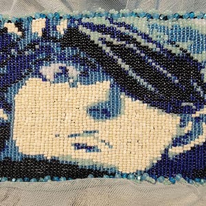 May include: A close-up of a beaded portrait of a person with blue and white beads. The person's hair is black and the background is blue and white.