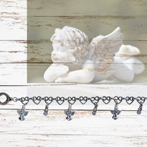 May include: Silver charm bracelet with heart-shaped links and angel wing charms. The bracelet has a lobster clasp closure and is shown on a white wooden background.