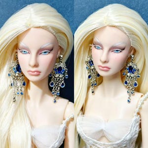 May include: A doll with long blonde hair and blue eyes wearing ornate silver-toned chandelier earrings featuring deep blue teardrop-shaped stones and clear crystals. The doll is dressed in a white textured garment with a pearl-beaded strap.