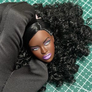May include: A doll head with dark skin, long black curly hair, and a side part. The doll features purple eyeshadow, white eyeliner, long eyelashes, and metallic purple lipstick. The facial expression is neutral with defined eyebrows and cheekbones.