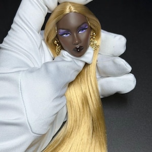 May include: A doll head with dark skin, long straight blonde hair, and purple eyeshadow. The doll features dark lips with a small white dot accent and wears gold-toned dangling earrings. The head is held by a white gloved hand.