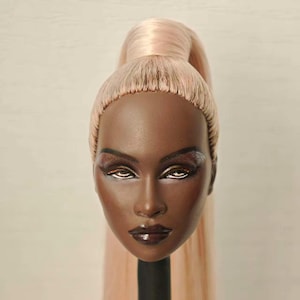 May include: A mannequin head with dark brown skin, styled with a high ponytail of light pink hair. The face features dramatic eye makeup with brown and silver eyeshadow, and dark lipstick. The head is mounted on a black stand.
