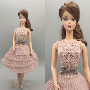 May include: A fashion doll wearing a pink dress with a lace bodice and a tiered skirt. The dress features a gray ribbon belt. The doll has brown hair styled in a ponytail and is wearing silver shoes. The doll is standing on a clear stand.