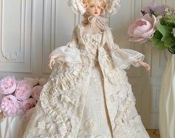 May include: A doll dressed in an elaborate cream-colored gown and matching hat. The dress features intricate lace, ruffles, and floral embellishments. The doll has blonde hair and is posed in front of a white wall with floral decorations.