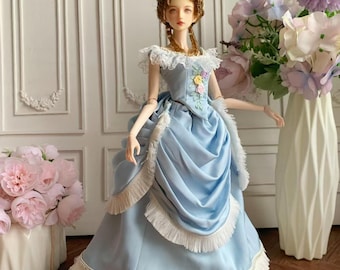 May include: A doll in a light blue gown with white fringe and floral accents. The doll has a brown wig and is posed in front of a white wall with decorative panels. A vase of flowers is to the right and a vase of pink flowers is to the left.