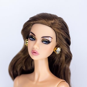 May include: A fashion doll with brown hair and dramatic eye makeup. The doll wears gold earrings with a clear stone center. The doll's skin tone is light, and the lips are a soft pink.