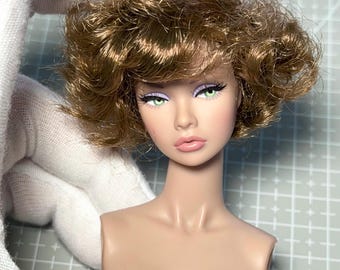 May include: A doll head with a fair complexion, green eyes, and full lips. The doll has a short, wavy, light brown hairstyle. The doll's face is made up with long black eyelashes and purple eyeshadow. The doll's neck and shoulders are visible.