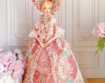 May include: A doll in a detailed, ornate gown and matching hat. The dress is primarily pink with white lace and blue accents. The doll has blonde hair and is posed in front of a white wall with decorative panels. The overall aesthetic is vintage and elegant.