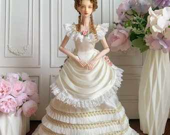 May include: A porcelain doll in a cream-colored gown with a layered skirt and gold trim. The doll has a high, elaborate hairstyle and is adorned with lace and small pink roses. The doll is posed in front of a white wall with decorative panels.