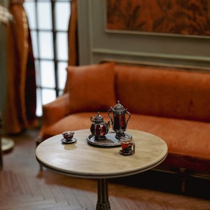 May include: A round wooden table with a silver teapot and two teacups filled with dark liquid. The table has a carved wooden base and sits on a wooden floor.