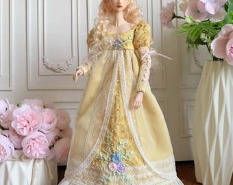 Puede incluir: Una muñeca con cabello rubio largo y ondulado que lleva un vestido amarillo con ribete de encaje blanco y bordado floral. El vestido tiene mangas largas y una falda amplia. La muñeca está de pie frente a una pared blanca con molduras decorativas, con flores rosas en el fondo.