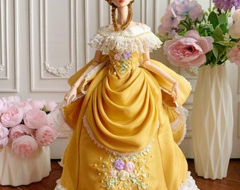 Puede incluir: Una muñeca detallada con un vestido amarillo mostaza con cuello de encaje blanco y falda con volantes. El vestido presenta detalles florales bordados en colores pastel. La muñeca tiene una peluca marrón peinada y posa frente a un arreglo floral.