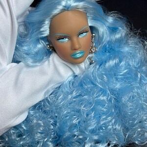 May include: A doll head with long, wavy, bright blue hair. The doll features tan skin, light blue eyeshadow, dark eyeliner, and glossy blue lips. It wears silver-toned dangling earrings with clear stones. The hair is voluminous and curly.