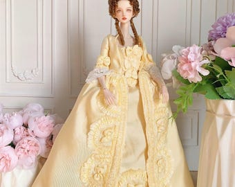 May include: A doll dressed in a yellow gown with detailed lace and bow embellishments. The doll has a brown wig styled in an elaborate updo with braids. The gown features a long coat with decorative trim. The background includes flowers.