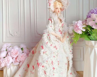May include: A doll in a white and pink floral gown and matching bonnet. The gown features intricate lace and ruffles, with a long train. The doll has blonde hair and is posed in front of a white wall with floral arrangements.