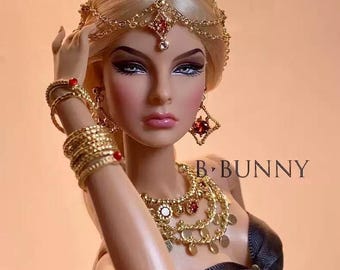 Può includere: Una bambola con capelli biondi e gioielli d'oro elaborati, tra cui un copricapo, orecchini, braccialetti e una collana con accenti rossi. La bambola indossa un top nero. Il testo "B. BUNNY" è visibile.