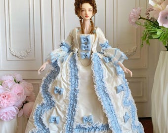 Puede incluir: Una muñeca con un vestido color crema con detalles en azul claro, lazos y adornos. El vestido presenta un diseño con volantes y una falda azul claro a juego. La muñeca tiene una peluca marrón peinada en un elaborado moño.