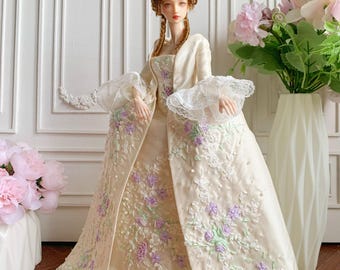 Puede incluir: Una muñeca detallada con un vestido color crema con intrincados bordados florales en morado y verde. La muñeca tiene un peinado alto y elaborado y lleva una chaqueta color crema a juego. El vestido presenta detalles de encaje y un dobladillo con volantes, creando una estética histórica y elegante.