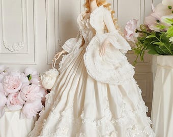 Puede incluir: Una muñeca de porcelana con un vestido de color marfil en capas con detalles de encaje y un sombrero a juego. La muñeca tiene el pelo rubio rizado y sostiene un pequeño bolso. El fondo presenta arreglos florales blancos y una pared blanca.