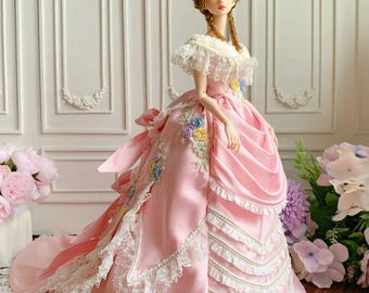 May include: A detailed doll in a pink satin gown with white lace trim and floral embroidery. The doll has a period hairstyle and is posed in front of a white wall with decorative molding. The dress features a layered skirt and a large bow.