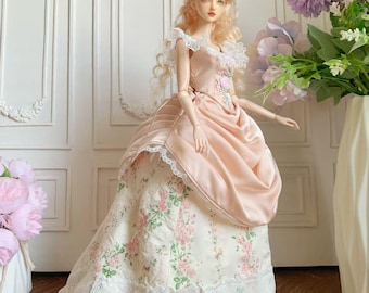May include: A doll wearing a pink and cream-colored ball gown with lace trim. The dress features a satin bodice and a floral patterned skirt. The doll has blonde hair and is posed in front of a white wall.