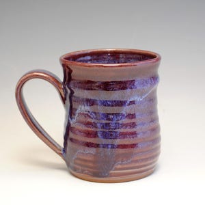May include: Handmade ceramic mug with a brown and purple glaze. The mug features horizontal bands of color and a curved handle. The mug is suitable for hot or cold beverages. The mug is approximately 4 inches tall.
