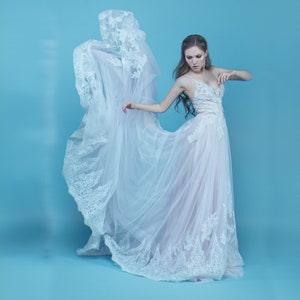 May include: A woman in a white lace wedding dress with a flowing skirt poses against a light blue background.