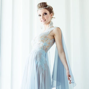 May include: A woman wearing a light blue, sheer, lace dress with a flowing cape. She is wearing a silver tiara and earrings. The dress has a fitted bodice and a flowing skirt.