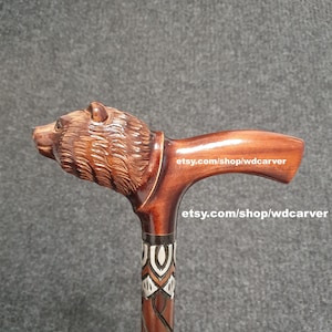 Pode incluir: Uma bengala de madeira com uma alça esculpida em forma de cabeça de urso. A bengala é marrom com um padrão decorativo em branco e preto. O texto "etsy.com/shop/wdcarver" está impresso na bengala.