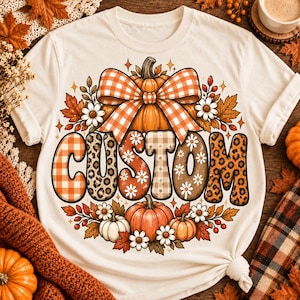 May include: A cream-colored t-shirt featuring a graphic of a large orange pumpkin topped with an orange and white checkered bow. Below the pumpkin, the word CUSTOM is printed in large letters with varied patterns including plaid, leopard print, and daisies.