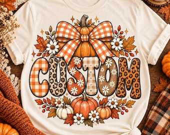 May include: A cream-colored t-shirt featuring a graphic of a large orange pumpkin topped with an orange and white checkered bow. Below the pumpkin, the word CUSTOM is printed in large letters with varied patterns including plaid, leopard print, and daisies.