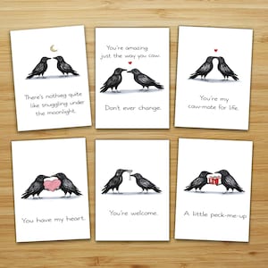 Puede incluir: Conjunto de seis tarjetas de felicitación con ilustraciones de cuervos negros y mensajes románticos. Las tarjetas tienen frases como "You're amazing just the way you caw" y "You have my heart". Las tarjetas están sobre una superficie de madera.