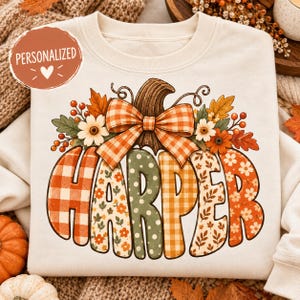May include: A cream-colored sweatshirt featuring a large graphic of a pumpkin with the name HARPER written across it. The pumpkin is decorated with a plaid bow, autumn leaves, berries, and flowers. A circular tag reads PERSONALIZED.