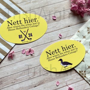 May include: Two yellow oval stickers with German text. One sticker has crossed hockey sticks and the numbers 81 and 24. The other sticker has a bird illustration. Both stickers have the text "Nett hier. Aber waren Sie schon mal in Shane Hollanders Cottage?" and "trishargh.com".