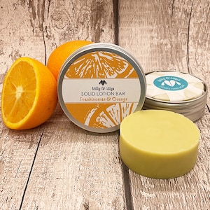May include: A round tin container with a white label that reads "Billy & Lilly's Solid Lotion Bar Frankincense & Orange". The tin is next to a round, yellow solid lotion bar and two orange halves.