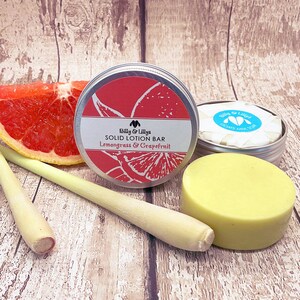May include: A round, yellow solid lotion bar in a silver tin with a red grapefruit and green lemongrass design. The tin has the text "Billy & Lilly's Solid Lotion Bar Lemongrass & Grapefruit". A grapefruit slice and two lemongrass stalks are in the background.