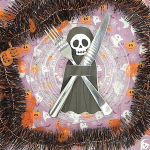 May include: A Halloween-themed table setting with a clear plate, a fork, and a knife. A black paper cutout of a grim reaper holds the silverware. The plate rests on a purple napkin with pumpkin and ghost designs, surrounded by orange and black garland.